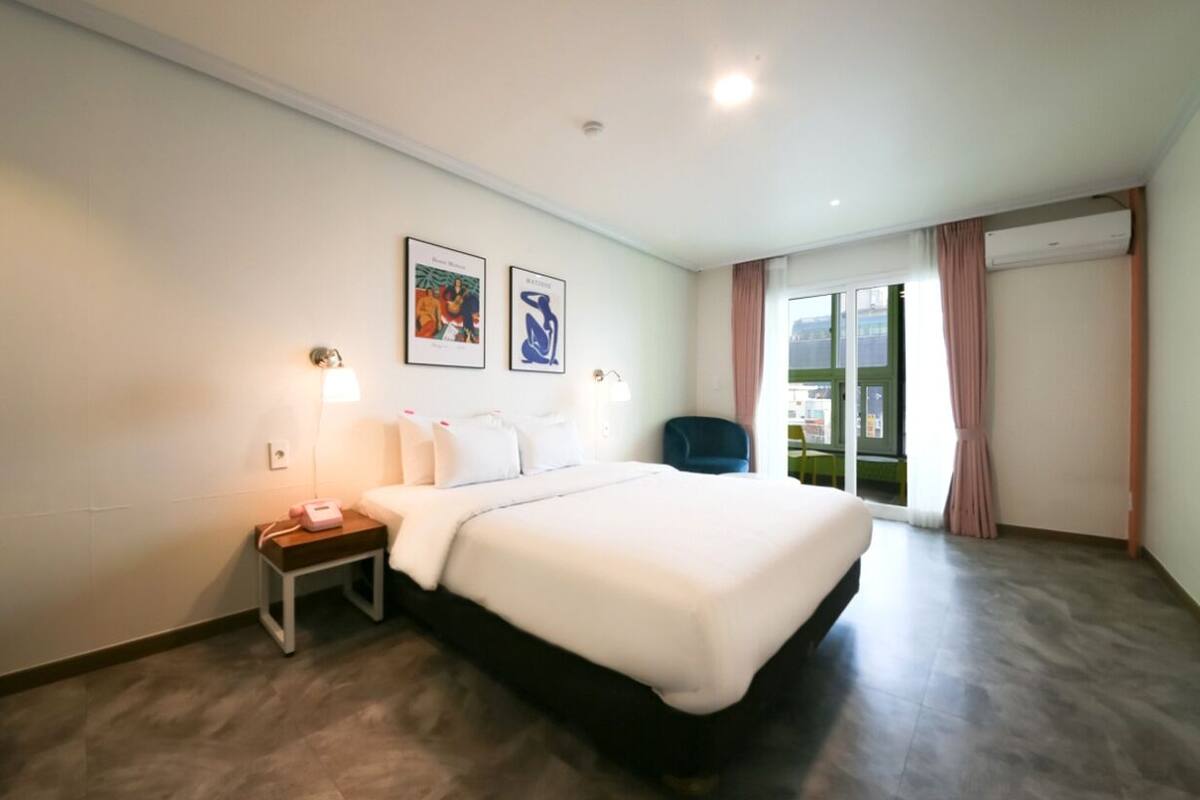 Une chambre d’hôtel avec un grand lit, une table de chevet avec une lampe, deux tableaux encadrés au mur et une vue sur l’extérieur par une fenêtre.