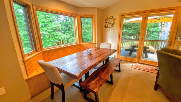 A dining area with a wooden table and benches, large windows with a view of greenery, and a sliding glass door leading to an outdoor deck. - O42 Renovated with high class upgrades! Ski access, boot warmer, ski rack, yard, AC, dog friendly! (Bretton Woods)