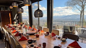 Un comedor con una mesa larga lista para una comida, ventanas amplias que ofrecen vistas a las montañas y lámparas colgantes.