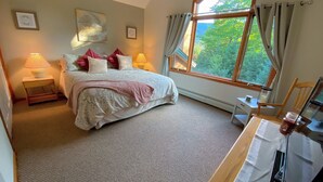 A bedroom with a large bed, two bedside lamps, a window with curtains, and a view of trees outside.