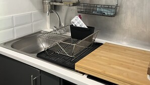 Eine Küche mit einer Spüle aus Edelstahl, einem schwarzen Geschirrgestell, einer Holz Schneidebrett und einem Metallregal mit Geschirrtuch.