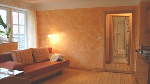 A cozy room with a wooden floor, a sofa with cushions, a table with a chair, a window with curtains, and a door leading to a bathroom. - House Froschkönig directly on the Weser (Hann. M ünden)