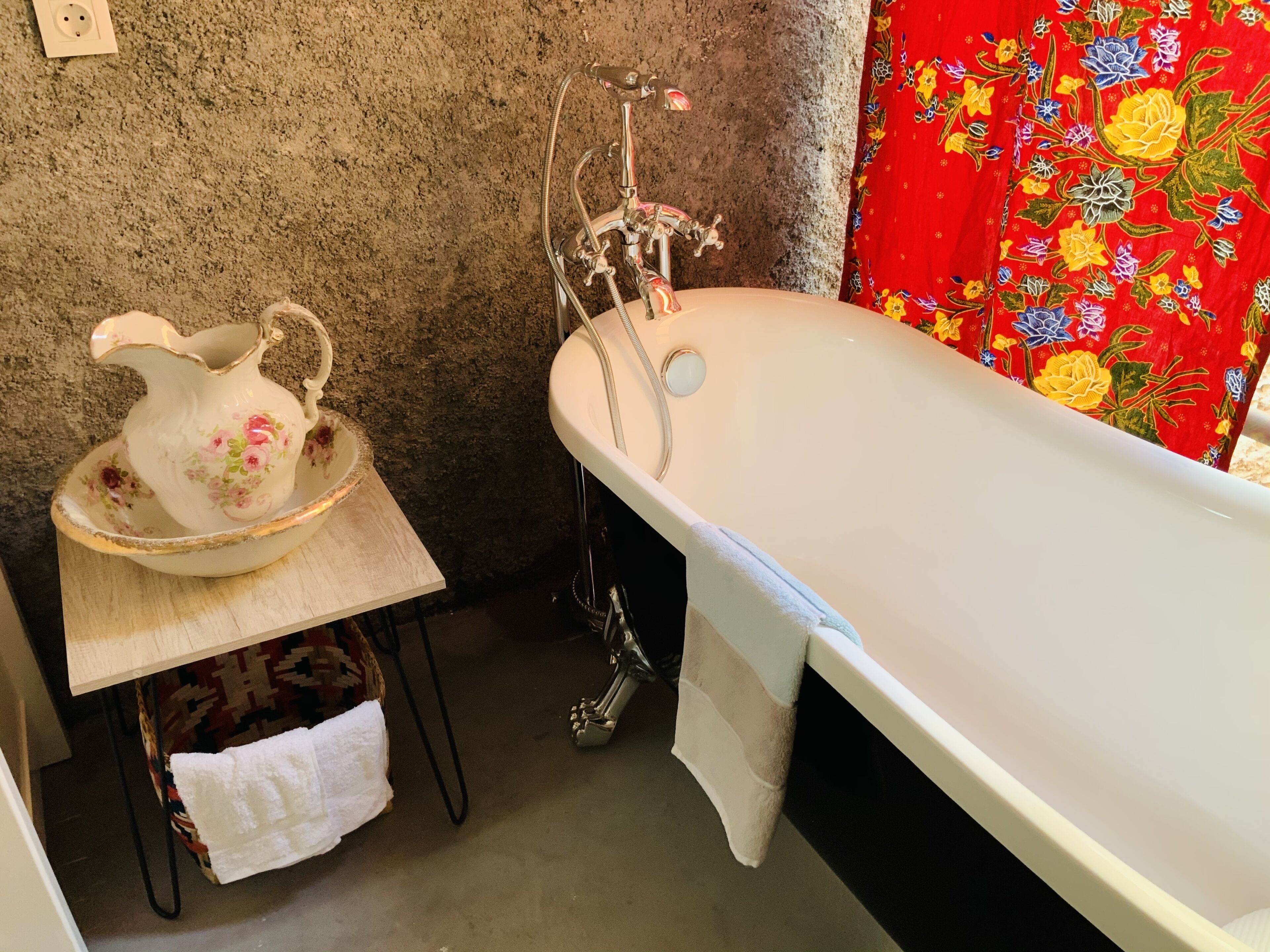 Un baño con una bañera, una jarra blanca sobre una mesa de madera y una cortina floral roja.