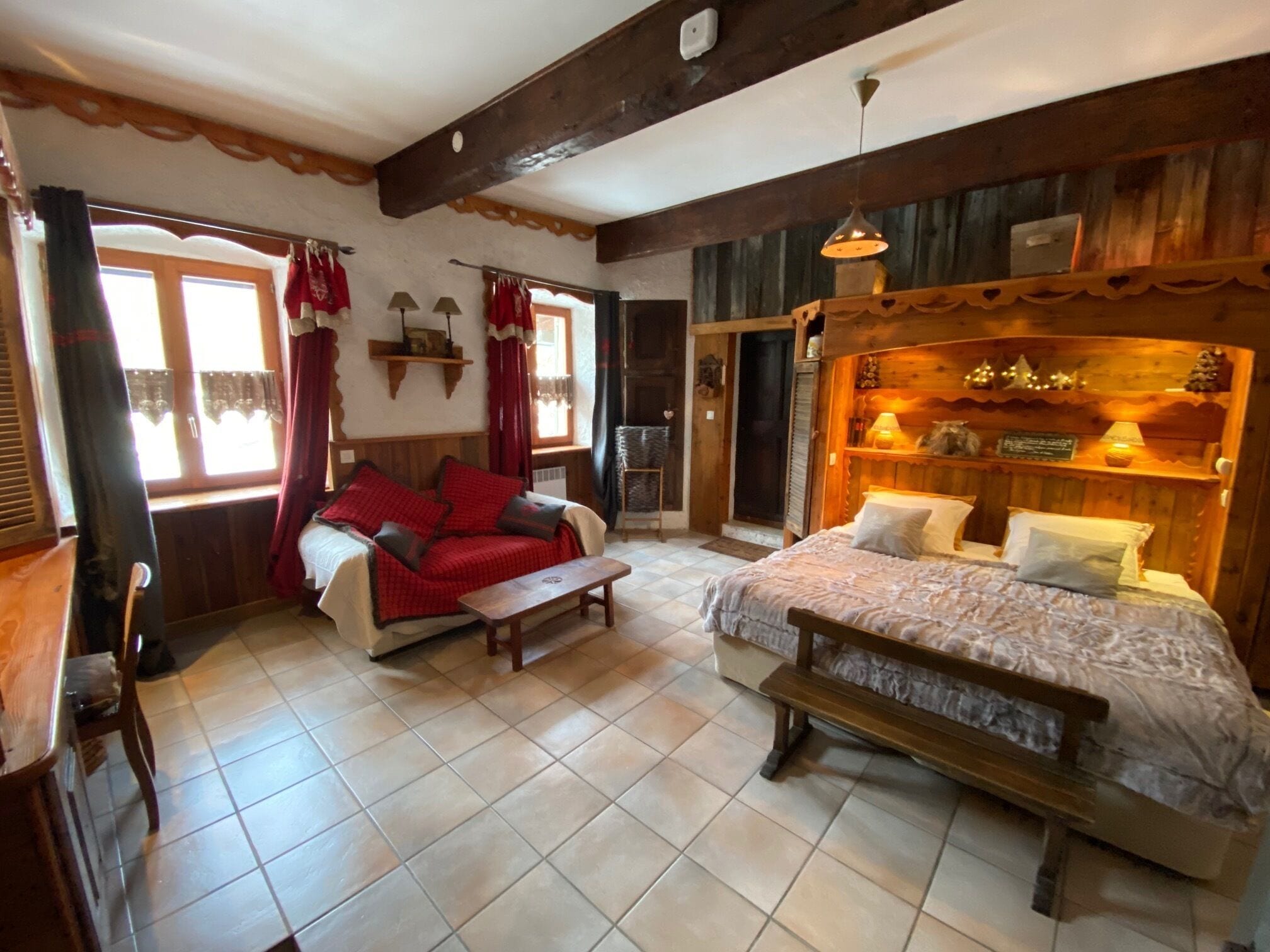A cozy room with a sofa, a bed, and a wooden bench. There are windows with curtains, a shelf with decorative items, and a wooden ceiling.