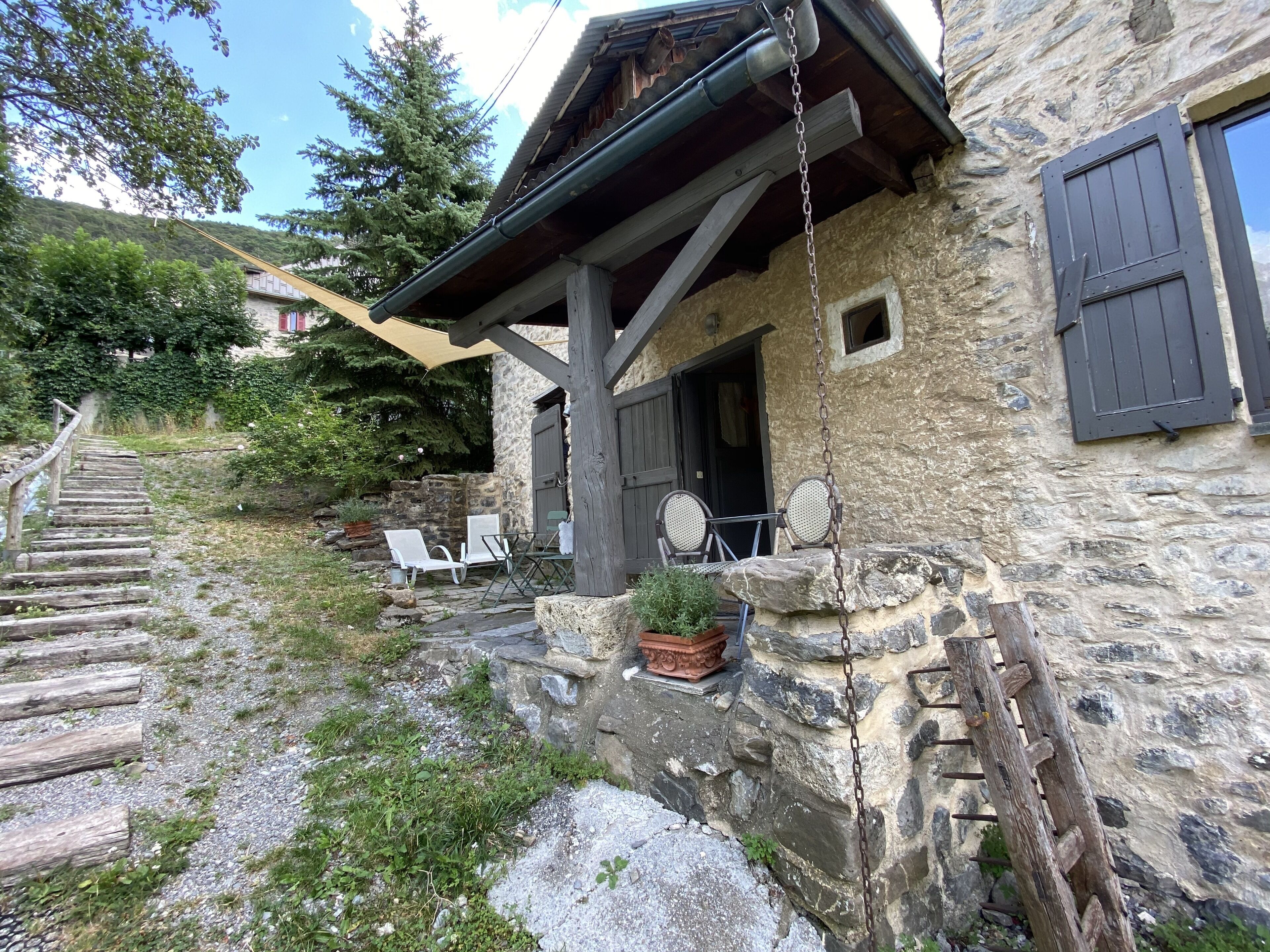 Ein rustikales Steinhaus mit einer Holztreppe, die zu einer überdachten Veranda führt. Unter einem Vordach stehen zwei weiße Stühle und ein Tisch.