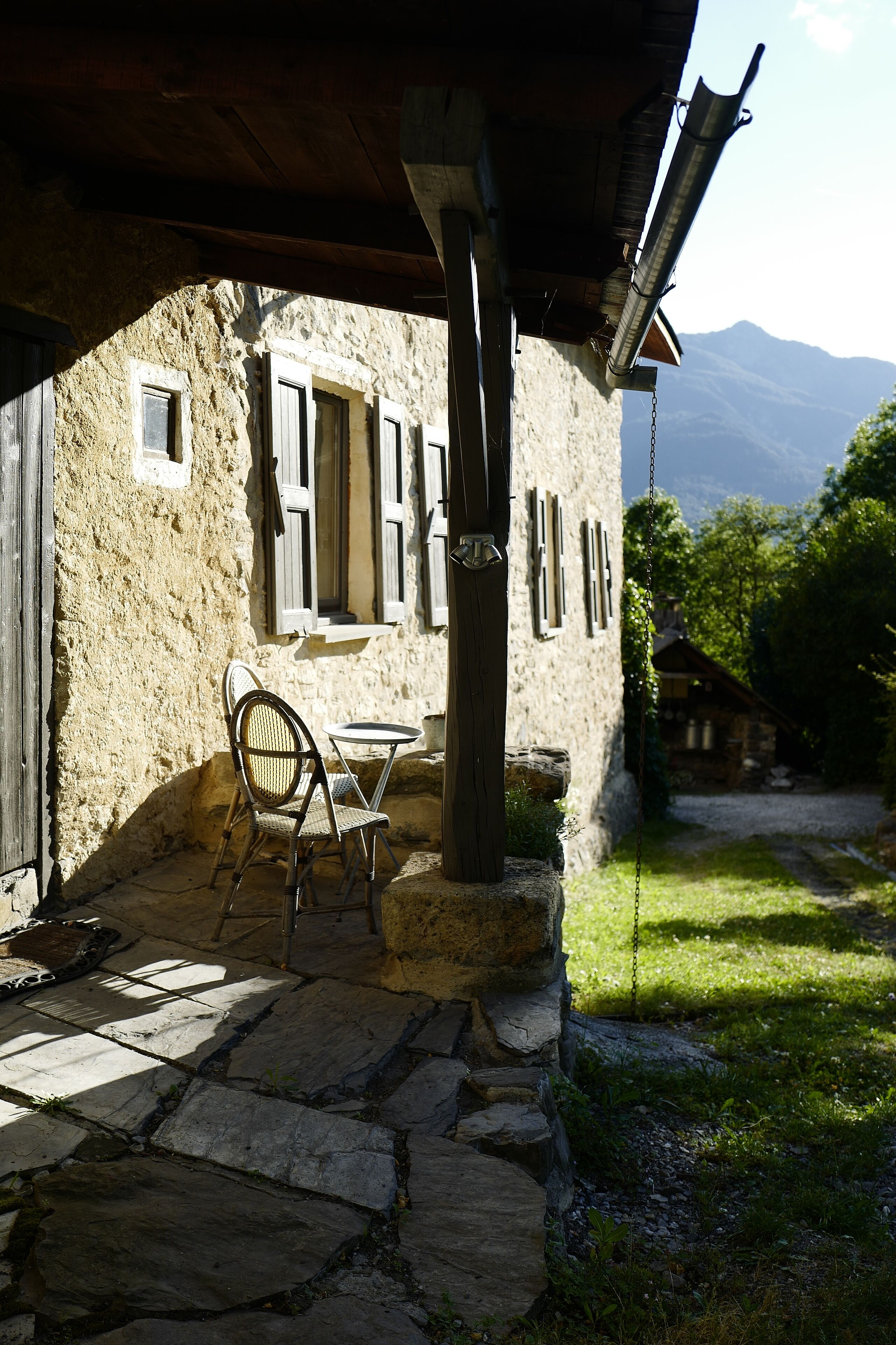 Un bâtiment rustique en pierre, doté d’un porche en bois, d’une table avec des chaises, et offrant une vue sur les montagnes en arrière-plan.