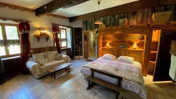 Una habitación acogedora con una cama, un sofá y una pequeña mesa. Tiene una ventana con cortinas, techo de madera y un estante con objetos.