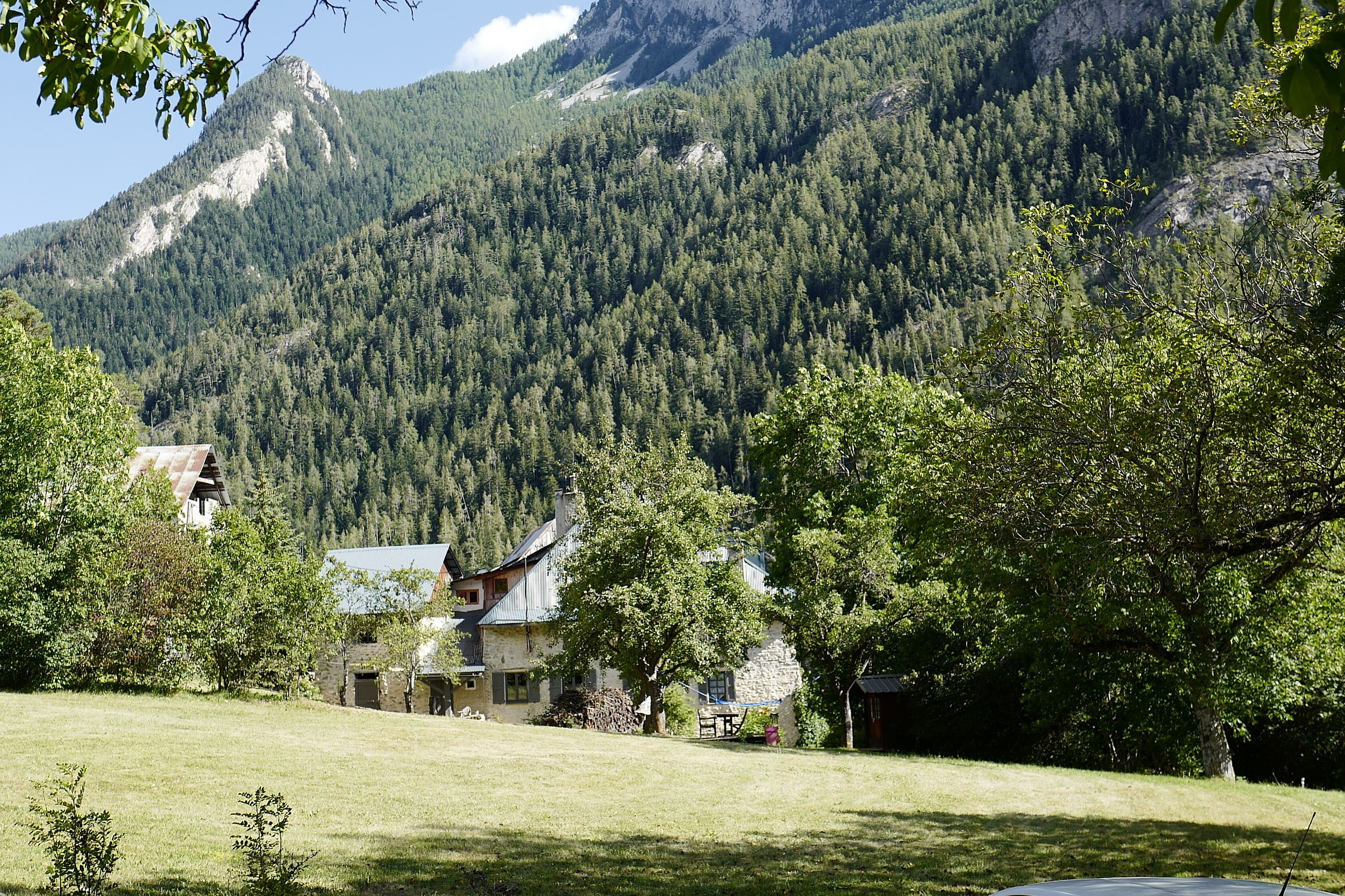 Ein rustikales Haus mit einer Stein-Fassade und einem geneigten Dach, umgeben von üppiger grüner Landschaft und einem bewaldeten Berg im Hintergrund.