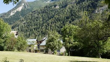Ein rustikales Haus mit einer Stein-Fassade und einem geneigten Dach, umgeben von üppiger grüner Landschaft und einem bewaldeten Berg im Hintergrund.