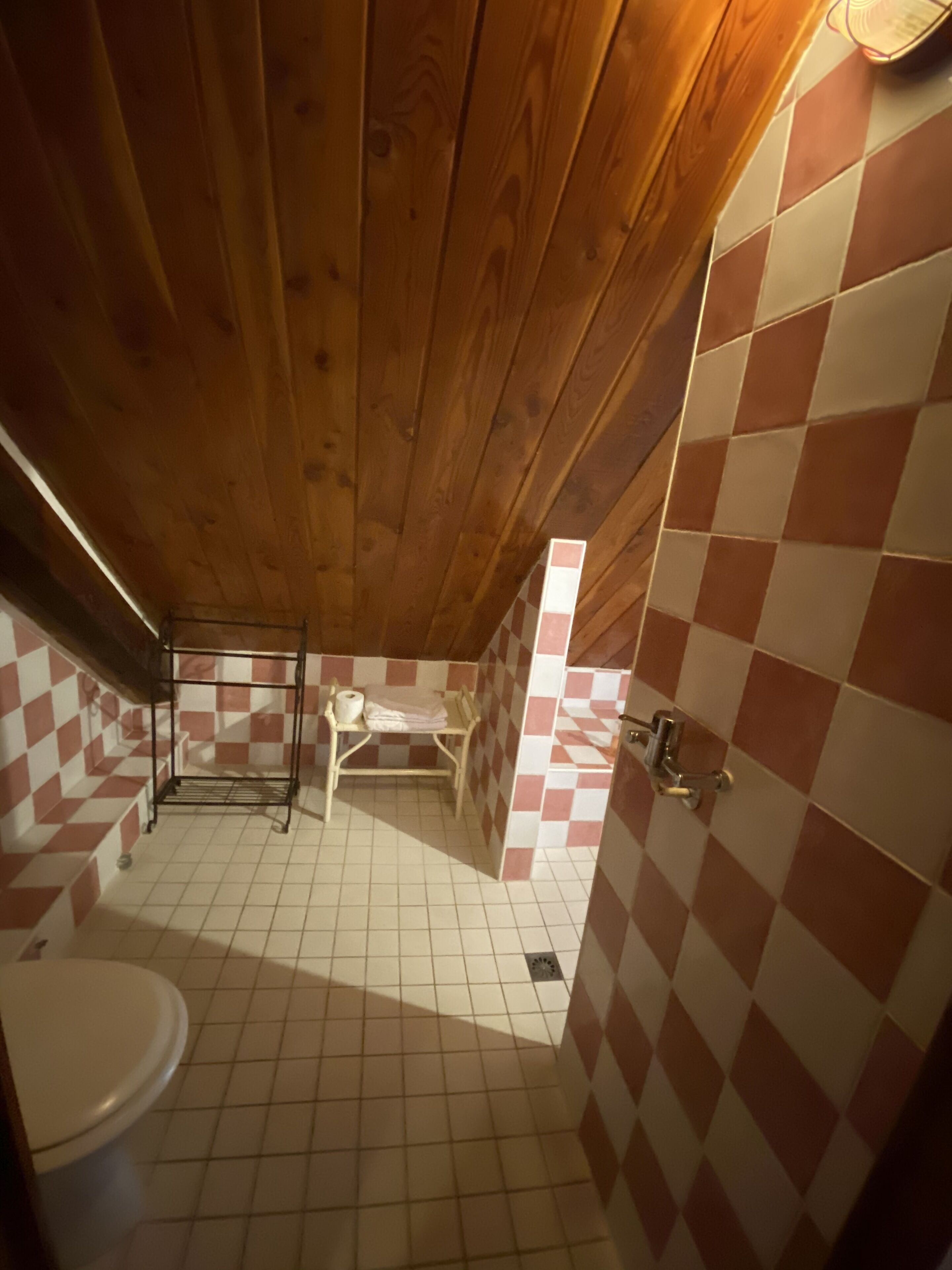 Un baño con techo de madera, paredes con baldosas de diseño de cuadros y un inodoro blanco, además de un perchero con toallas.