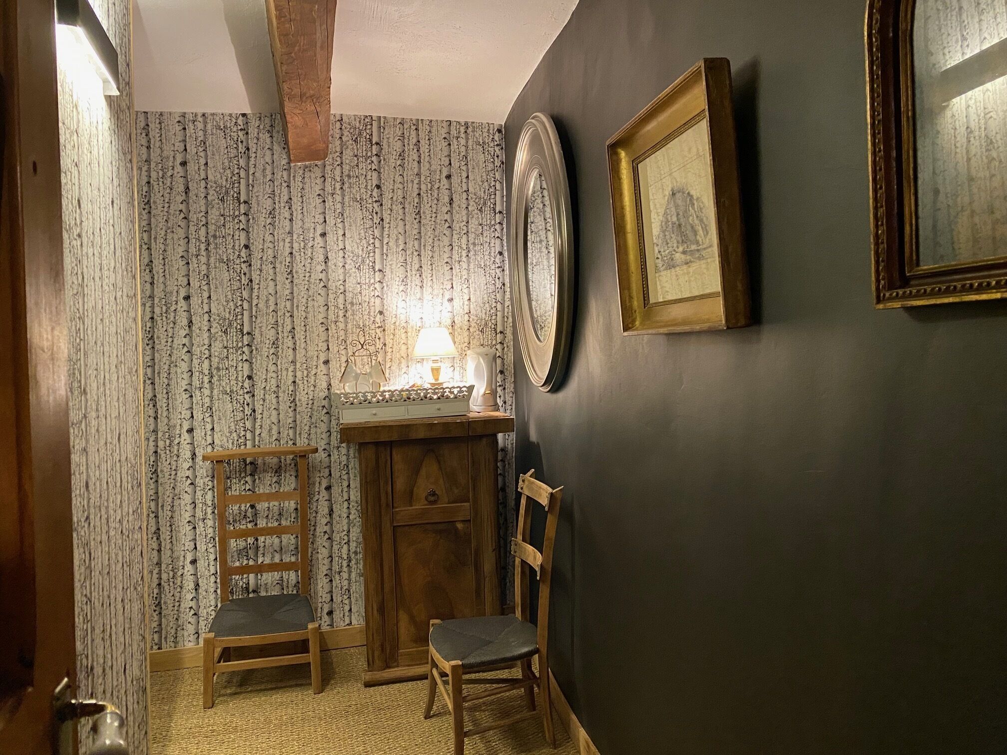 Una habitación con pared oscura, un armario de madera, dos sillas, una lámpara y cuadros enmarcados.