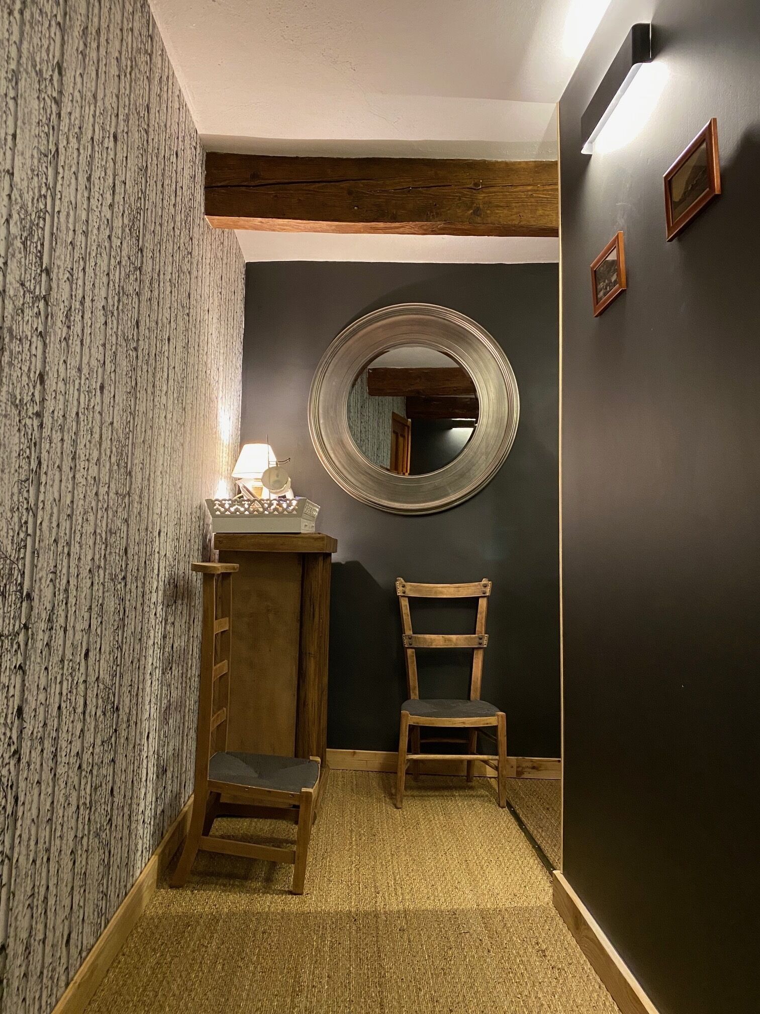 Un corridoio stretto con una sedia di legno, un tavolino con una lampada e uno specchio rotondo appeso al muro.