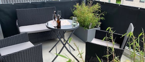 Un balcón con mobiliario de mimbre, una mesa con bebidas y una planta en maceta.