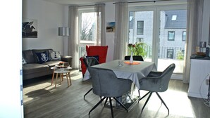 A modern living room with a grey sofa, a red armchair, and a dining table with grey chairs. There is a window with curtains, a plant, and a view of buildings outside.