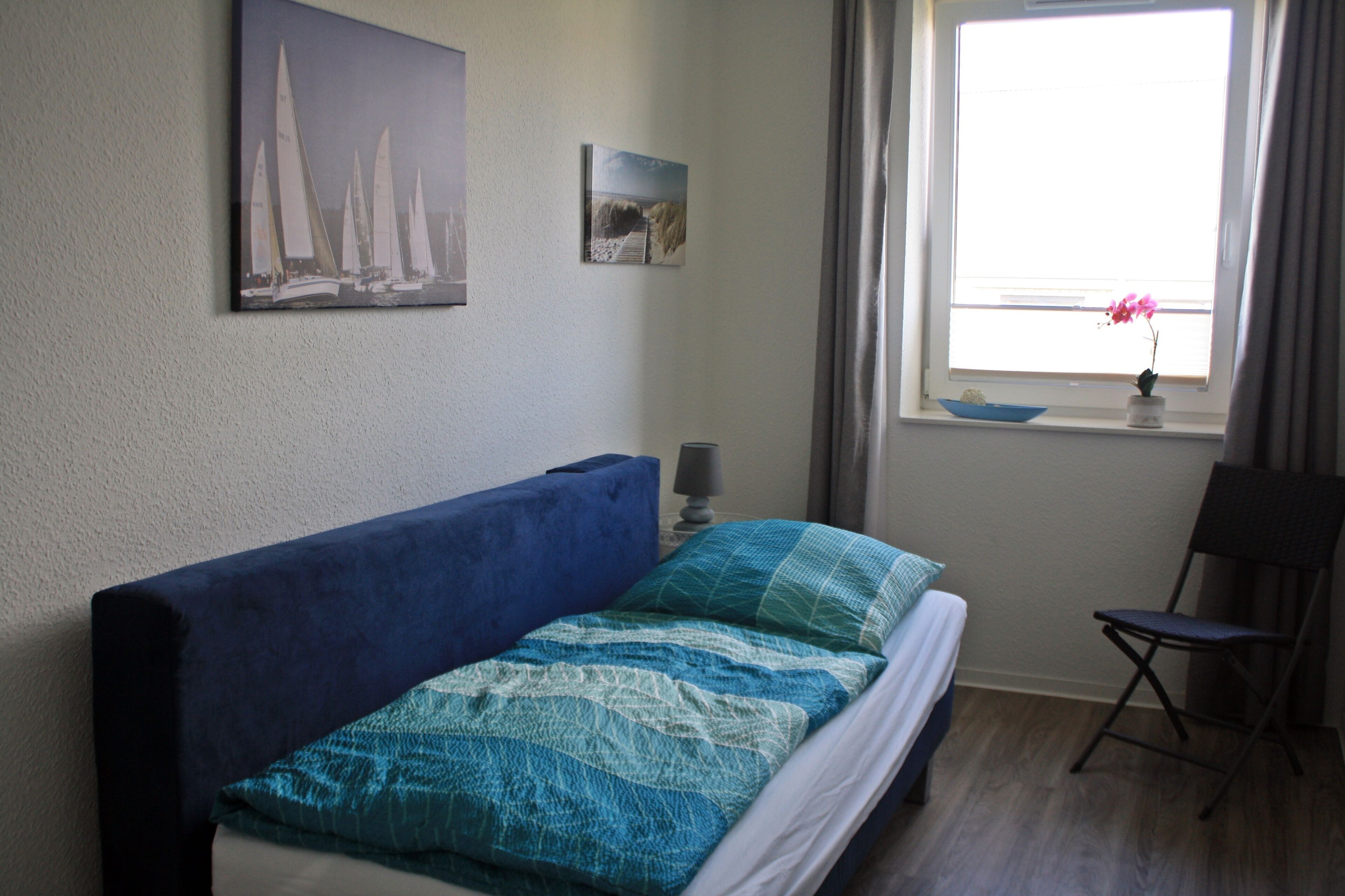 Un dormitorio con una cama, una silla, un ventanal con cortinas y dos cuadros colgados en la pared.