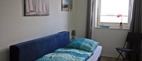 Un dormitorio con una cama, una silla, un ventanal con cortinas y dos cuadros colgados en la pared.