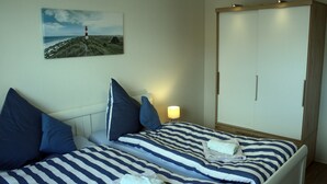 A bedroom with a bed featuring striped bedding, a nightstand with a lamp, a wardrobe, and a painting of a lighthouse on the wall.