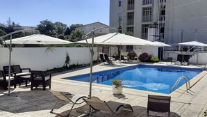 Una piscina con lettini e ombrelloni, circondata da un muro bianco e un edificio sullo sfondo.