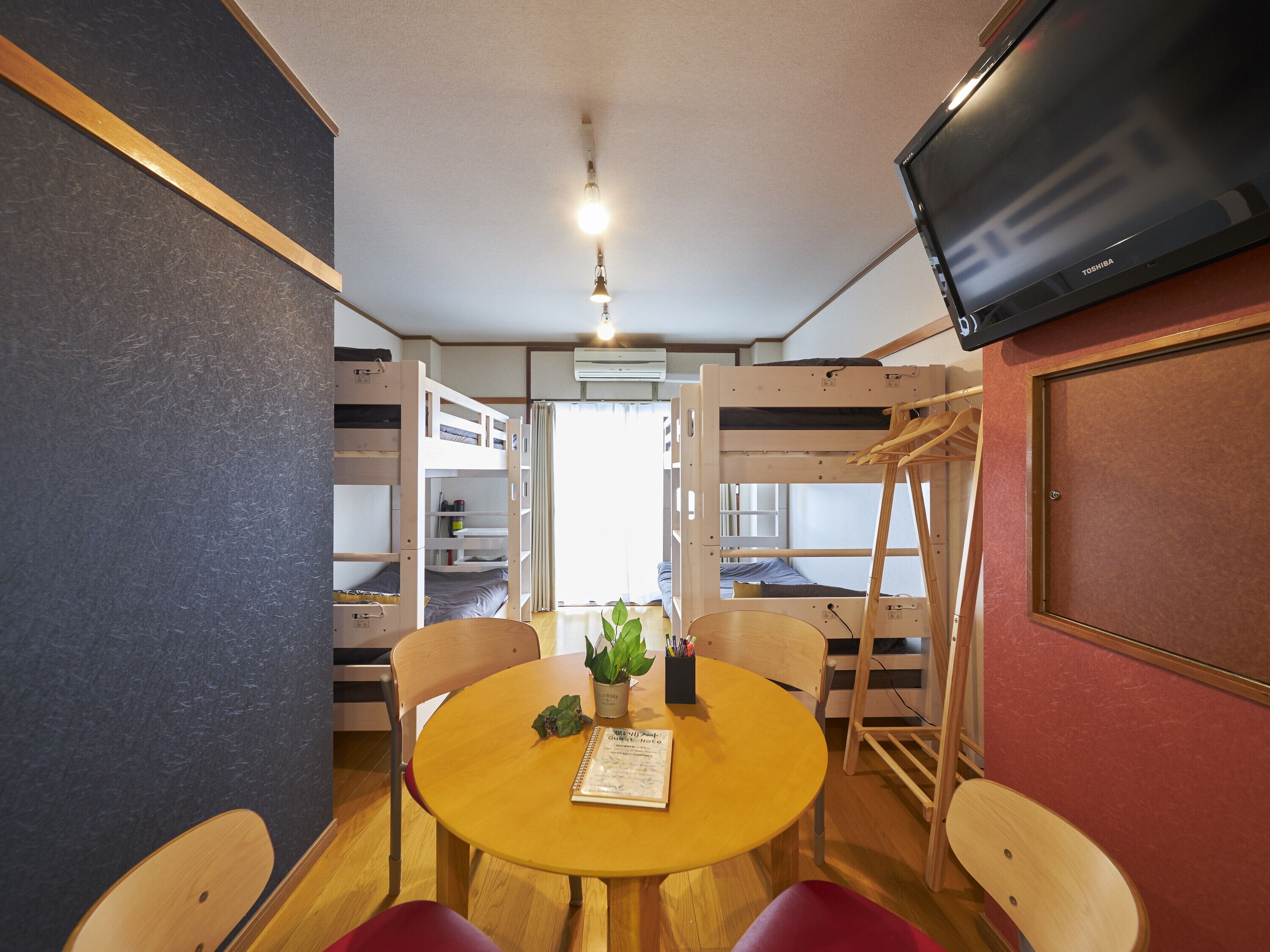 A room with bunk beds, a round wooden table with chairs, a potted plant, and a television on the wall.