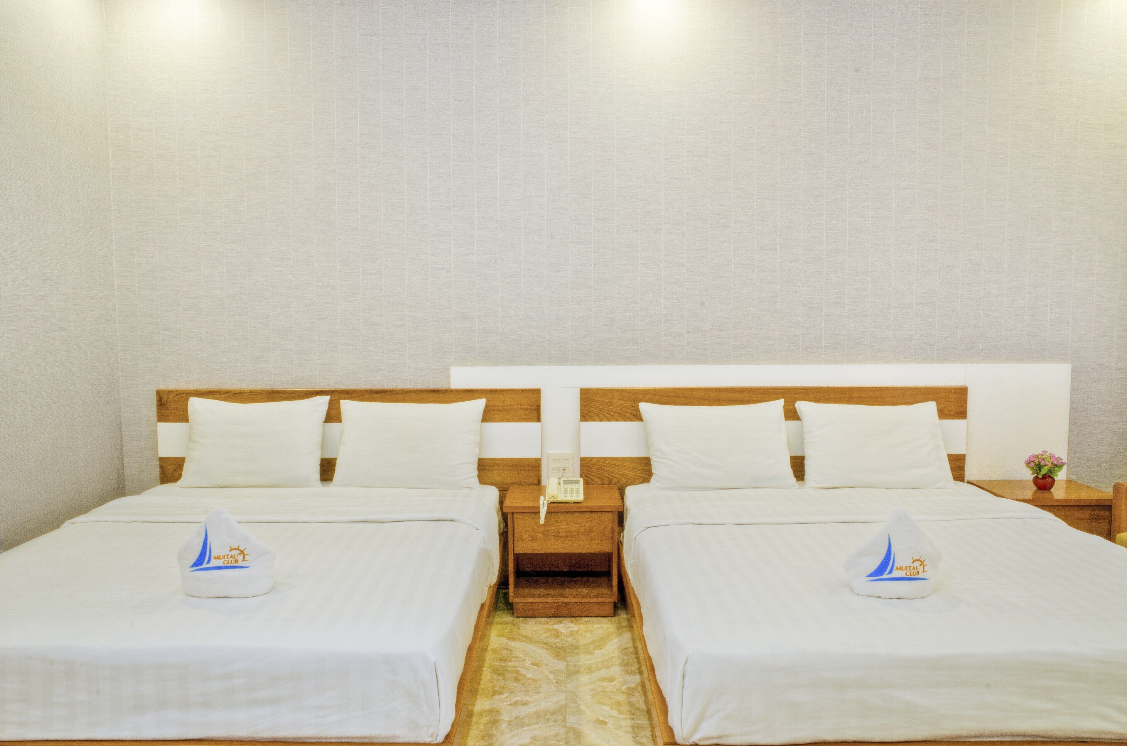 Habitación de hotel con dos camas, cabeceros de madera, ropa de cama blanca, una mesita de noche con un teléfono y un florero con flores.