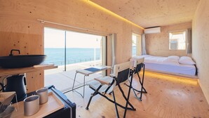 A compact, wooden cabin-style room with a bed, a small table with kitchen items, a chair, and a view of the sea through large windows. - Snow Peak Osaka Rinku (Izumisano)