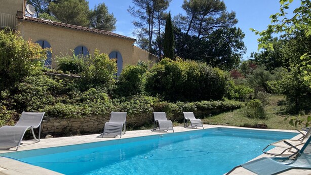 A rectangular swimming pool with clear blue water, surrounded by stone walls and greenery. There are four lounge chairs along the poolside.