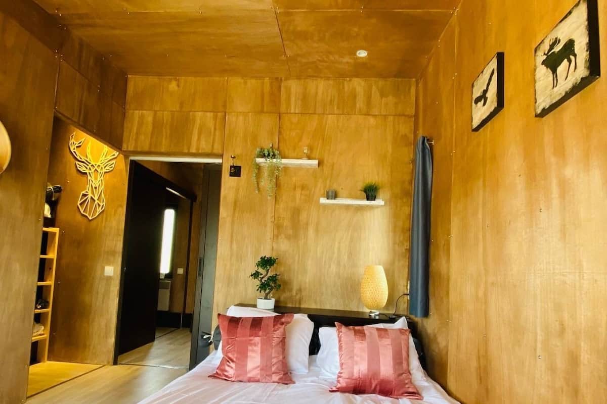 A bedroom with a wooden ceiling, a bed with white and pink bedding, a bedside table with a lamp, and a wall-mounted shelf with plants.