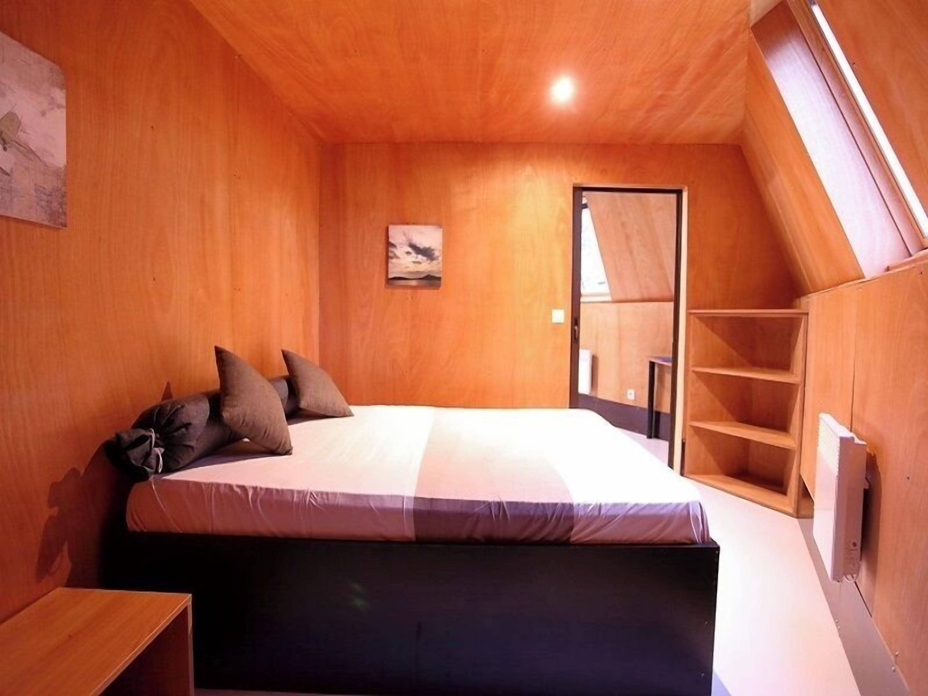 Ein Schlafzimmer mit Bett, Holzwänden, einem kleinen Schreibtisch und einer geneigten Decke.
