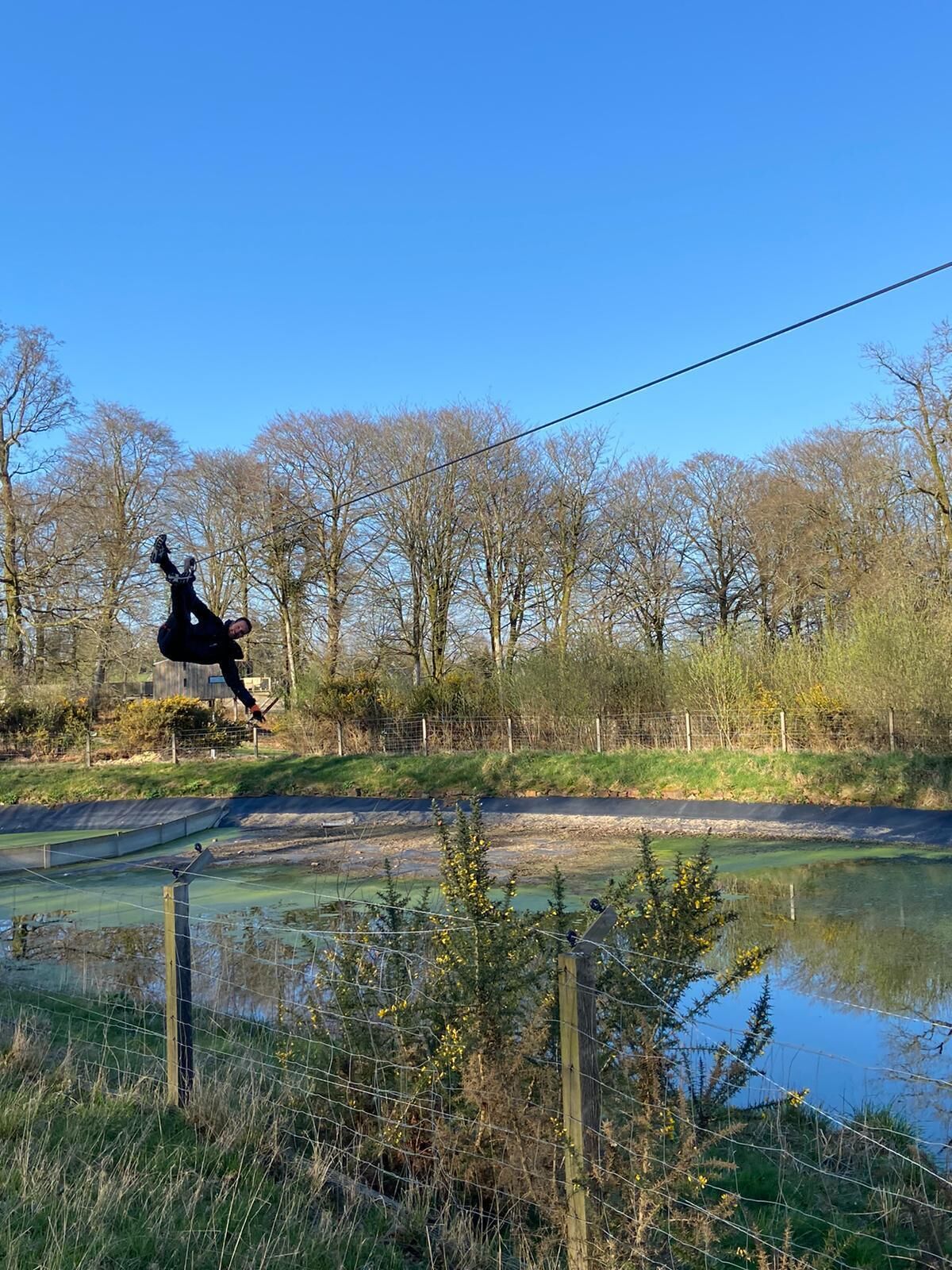 A person performing a high jump on a trampoline in a natural setting with trees and a pond.