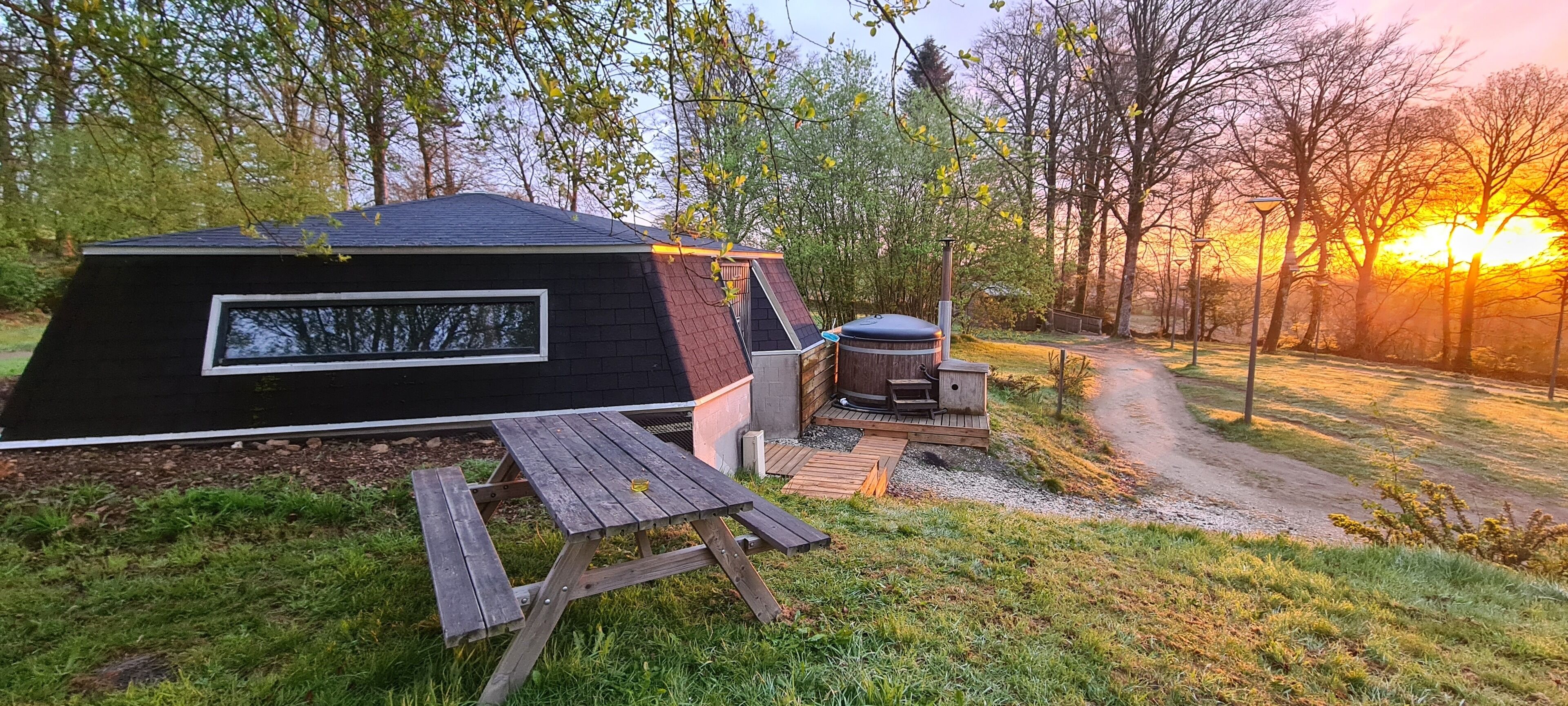 A modern, black-roofed cabin with a large window, a hot tub, and a picnic table in a wooded area.