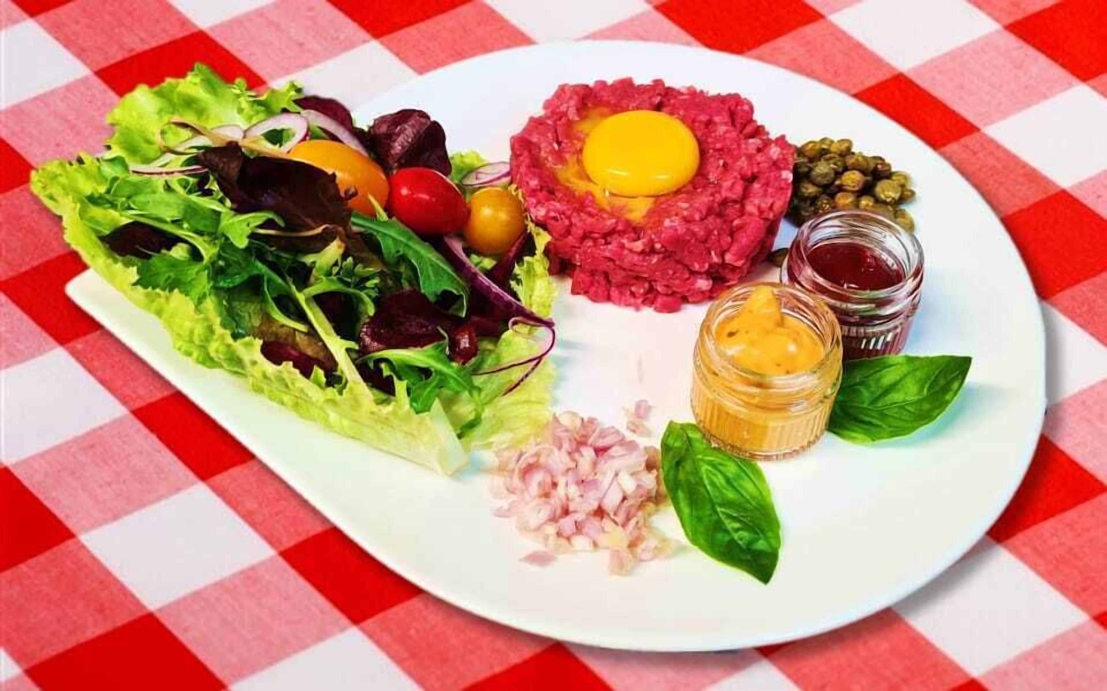 Une assiette comprenant une salade, un jaune d’œuf cru posé sur une base de viande, des cornichons et diverses sauces.