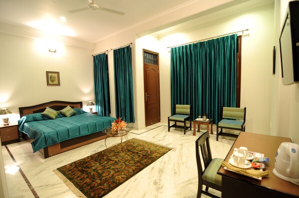 A hotel room with a bed, two chairs, a table with a kettle, and a small table with a lamp.