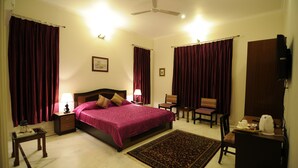 A hotel room with a bed, a desk, a chair, a table, a ceiling fan, and curtains. - Geetanjali Luxury Suites (Jaipur)