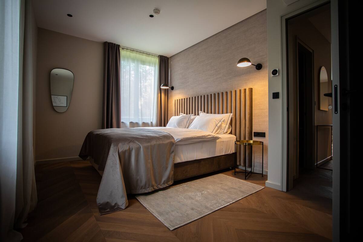 A modern bedroom with a bed, bedside tables, a wall-mounted light fixture, and a window with curtains.