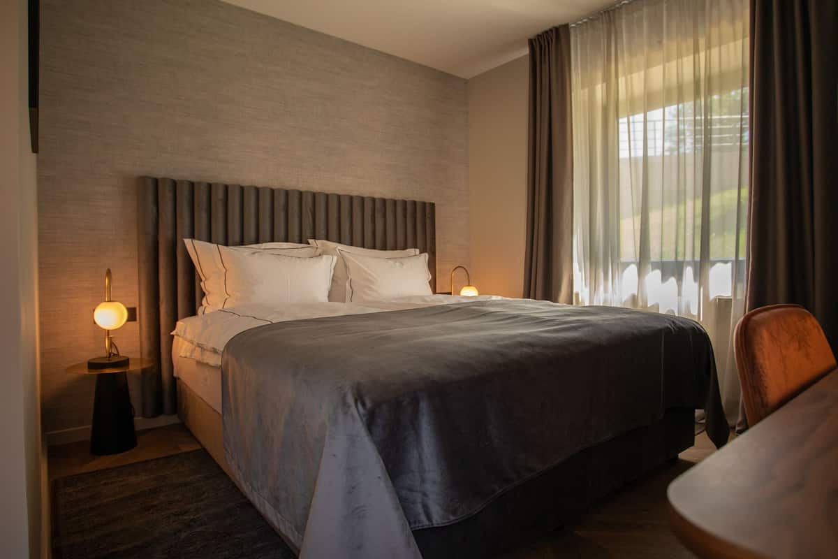 A modern hotel room with a large bed, two bedside tables with lamps, a wooden chair, and a view of greenery through the window.