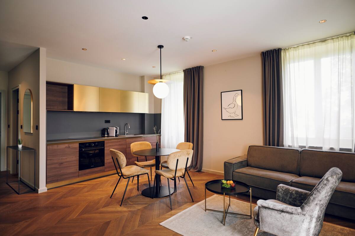 A modern living room with a sofa, a dining table with chairs, a kitchen area, and a large window with curtains.