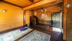 Una habitación amplia con paredes de madera y un techo alto. Se observa una puerta que da a otra habitación, una pequeña escalera y un tapete grande en el suelo.