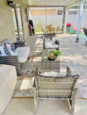 Un patio al aire libre con mobiliario de mimbre, incluyendo sofás y sillones, una mesa con una planta y una sección cubierta con vista al jardín trasero.