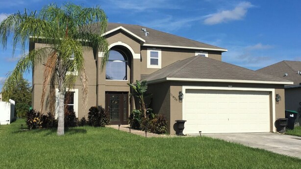 A two-story house with a garage, a large arched window, and a palm tree in the front yard. - Mi Casa Es Su Casa!!🏡 4 Bdrm home with pool (Orlando)