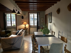Una sala de estar acogedora con un sofá, una mesa de comedor y una lámpara. Hay ventanas con cortinas, un radiador y una planta sobre la mesa.