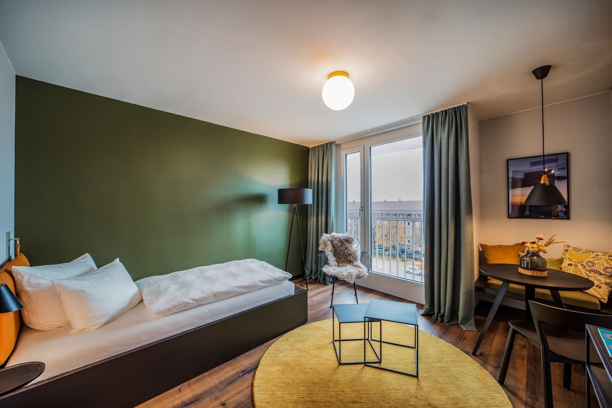 A modern hotel room with a bed, a round table, a chair, a sofa, a balcony view, and a hanging light fixture.