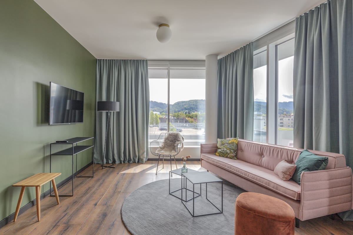 A modern living room with a sofa, a coffee table, a TV, and a view of a cityscape through large windows.