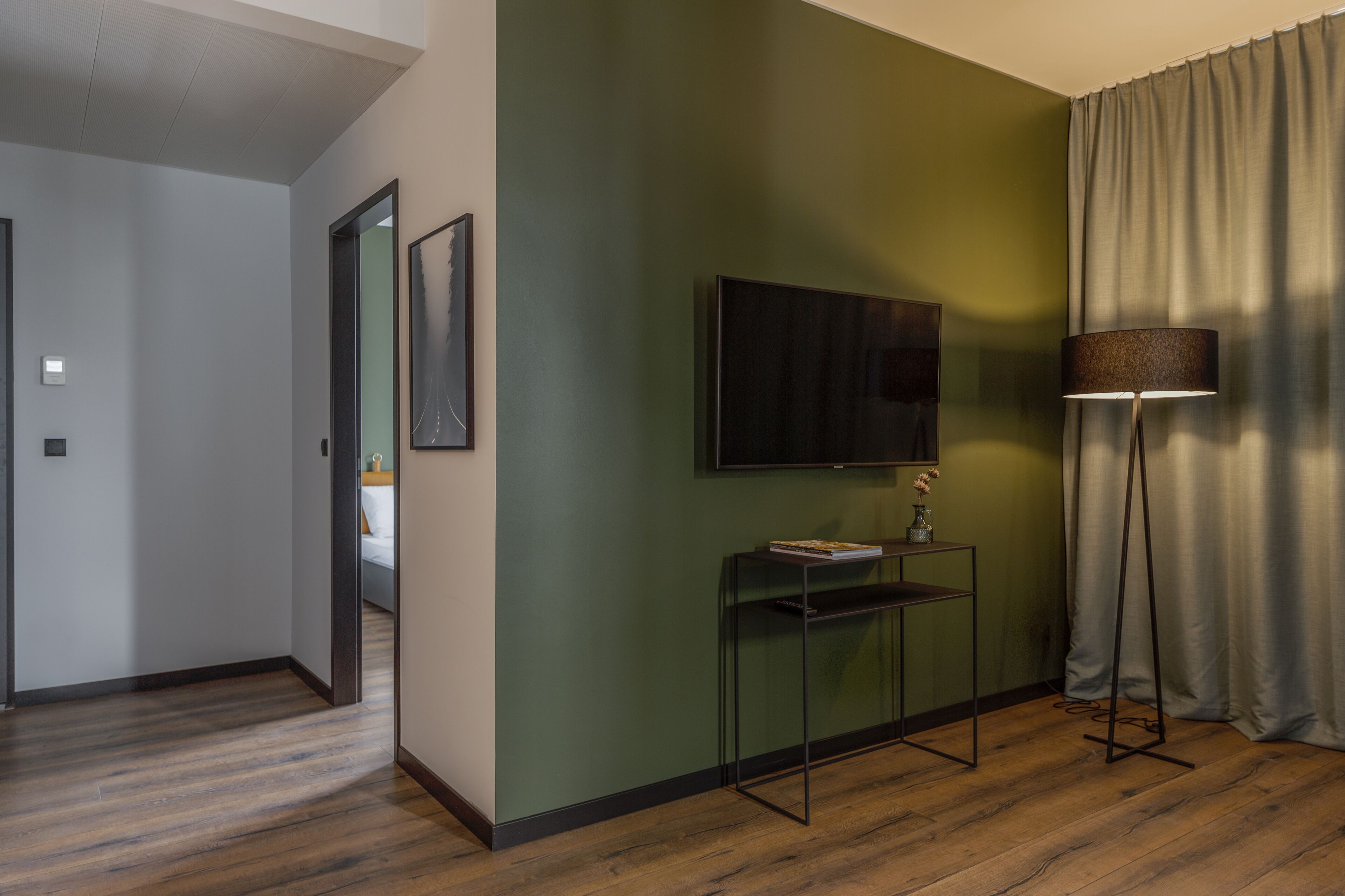 A room with a wooden floor, a green wall, a flat-screen TV mounted above a small table, and a floor lamp.