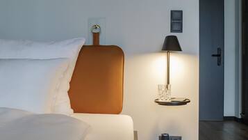 A neatly made bed with white linens, a brown headboard, a bedside lamp, and a small shelf with a glass.