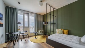 Une chambre moderne avec un lit, un bureau, des chaises et une vue sur la ville grâce à de grandes fenêtres.