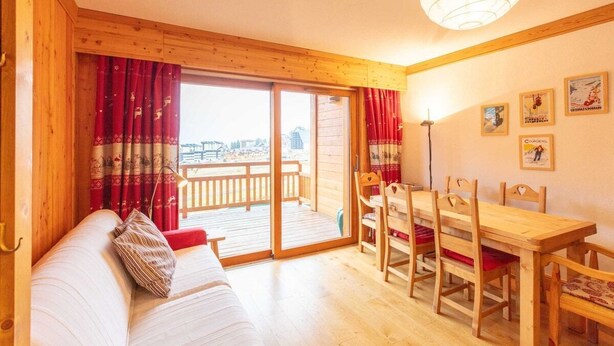 A cozy room with a sofa, wooden dining table, chairs, and a balcony with red curtains.