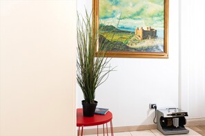 A room with a painting of a castle, a potted plant, a red stool, and a vacuum cleaner.