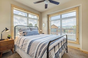 A bedroom with a bed, nightstand, lamp, and windows. - Edgewater Cabin on the Fraser River | 3BR Sleeps 7 | Kids Trout Pond & Pool (Granby)