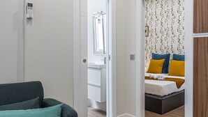Una habitación moderna y funcional con un sofá gris, suelo de madera, cama con almohadas amarillas y azules, y pared blanca con ventana.