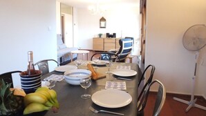 A dining area with a table set for four, including plates, cutlery, and a bottle. There are chairs around the table, a fan, and a small cabinet in the background. - Collioure Town with Parking – Apartment Sorra (Collioure)